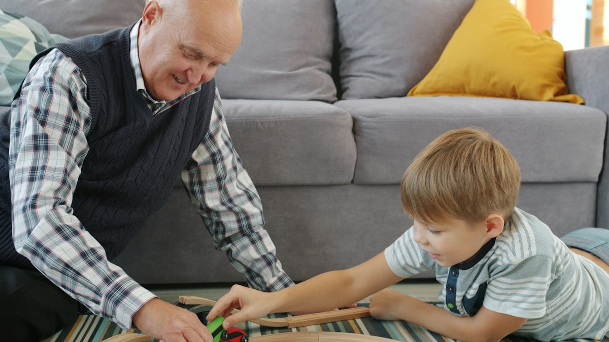 Happy boy is playing toy cars with loving grandad moving autos together smiling sitting on floor in apartment. Family relationship and lifestyle concept.
